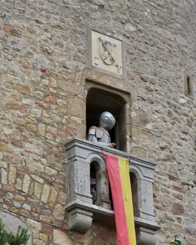 Outside view of Museum of History in Le Mont Saint Michel: wall of medieval building, window with armored knight figure and red - yellow flag